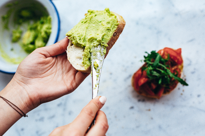 Receitas de abacate. Mão feminina está segurando uma fatia de pão enquanto passa a pasta de guacamole, feita com abacate, no pão. Na mesa de mármore branco tem uma tigela com a receita que leva abacate e outra fatia de pão com tomates vermelhos decorando.