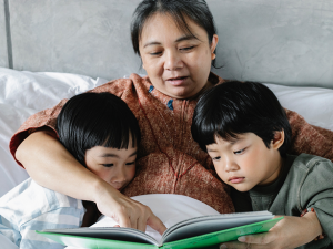 Leitura na infância. Mulher e duas crianças estão sentadas na cama. Eles possuem o cabelo liso e preto. A mulher está lendo um livro para as crianças e apontando para a folha.