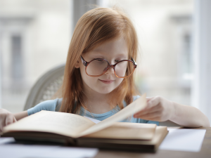 Leitura na infância. Menina branca de cabelos ruivos, usa óculos redondo e está sentada enquanto folheia um livro