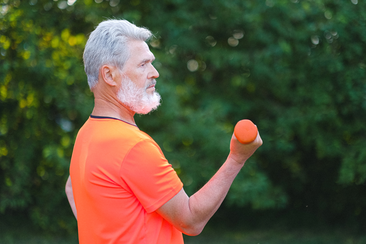 Atividade física em casa. Homem de pele branca, cabelos e barba grisalhos, aparenta estar na faixa dos 50 anos de idade. Ele veste uma camisa laranja e segura na mão direita um peso de ginástica também de cor laranja. Ele está em um lugar ao ar livre que tem árvores ao fundo.