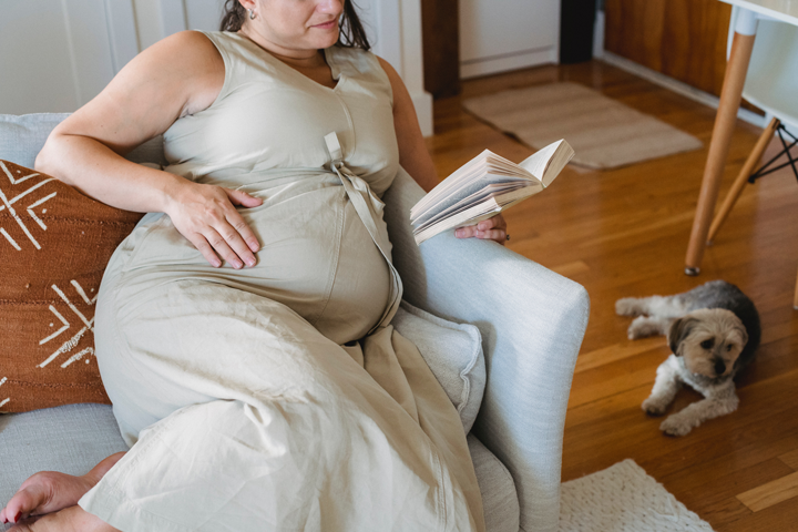 Para ser uma rede de apoio a gestantes e puérperas é preciso contribuir para a qualidade de vida da mãe, facilitando que ela tenha tempo disponível para realizar atividades que lhe dão prazer.Mulher grávida está sentada no sofá. Ela usa um vestido de tons claros, na mão esquerda está segurando um livro e a mão direita está apoiada na barriga. Atrás dela tem um cachorro de porte médio deitado no chão de madeira.