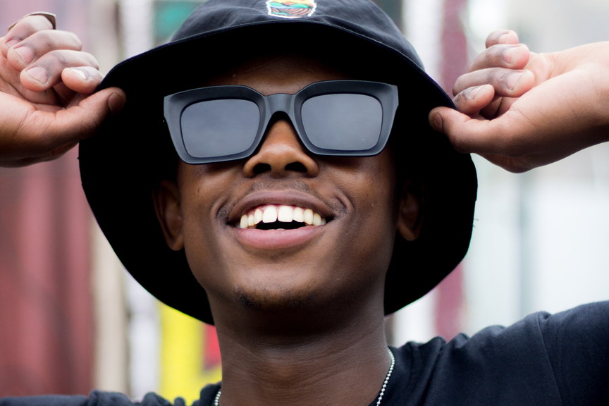 Homem negro usa óculos escuros. Ele está sorrindo enquanto segura o chapéu que está em sua cabeça. O chapéu é preto e a armação do óculos também é de cor preta.