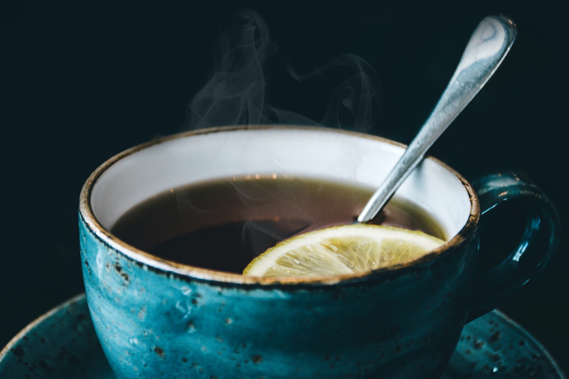 Xícara de cor azul esverdeada contém chá que traz benefícios para a saúde. No chá natural tem uma rodela de limão e há uma colher. O chá está quente e há fumaça saindo da xícara. 