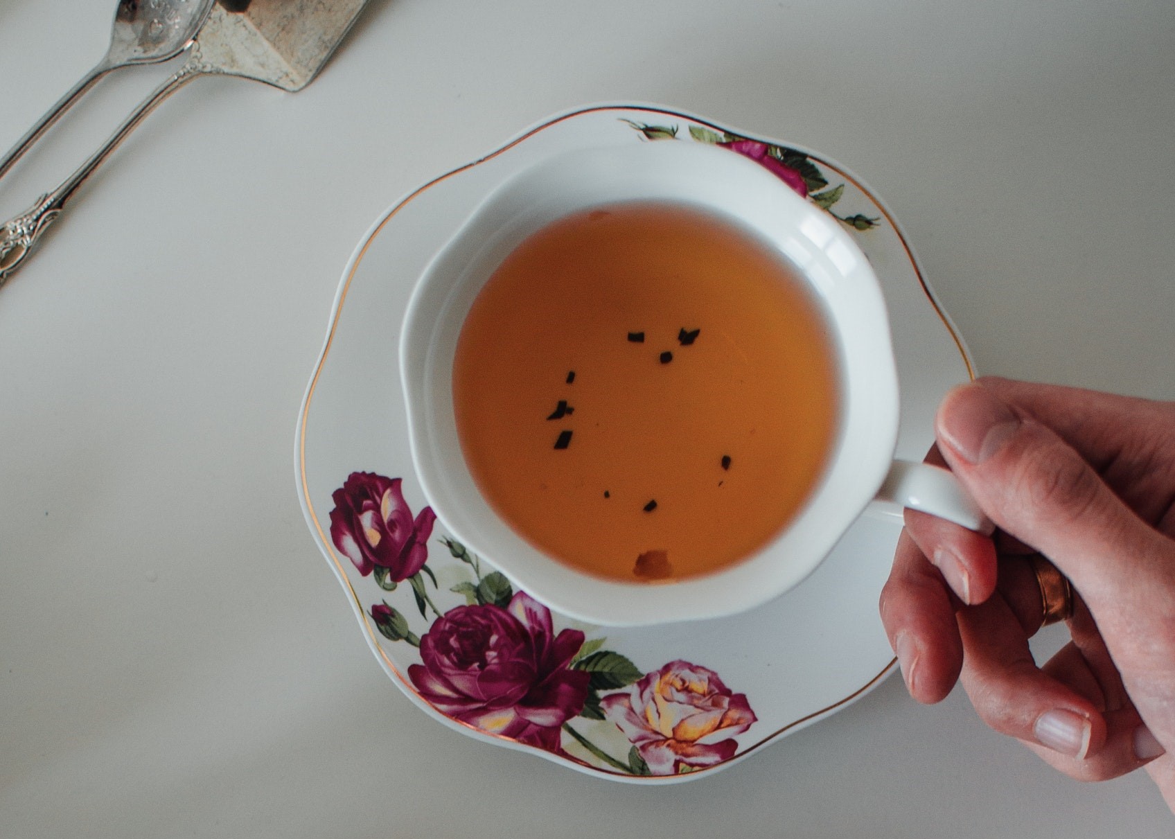 Mão de mulher segura uma xícara de chá. A xícara é de cor branca e está sobre um pirex que também é branco e possui decoração com rosas. 