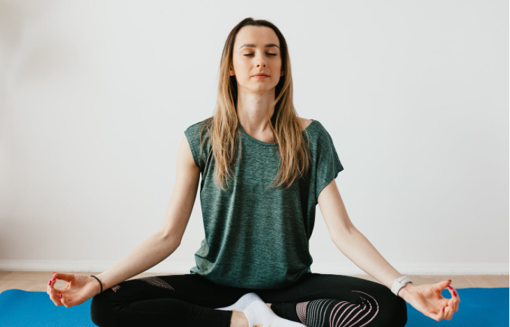 Mulher branca de cabelos loiros está praticando yoga para relaxar. Ela veste roupas adequadas para o exercício, camisa de cor verde e calça preta. Ela está sentada com as pernas cruzadas.