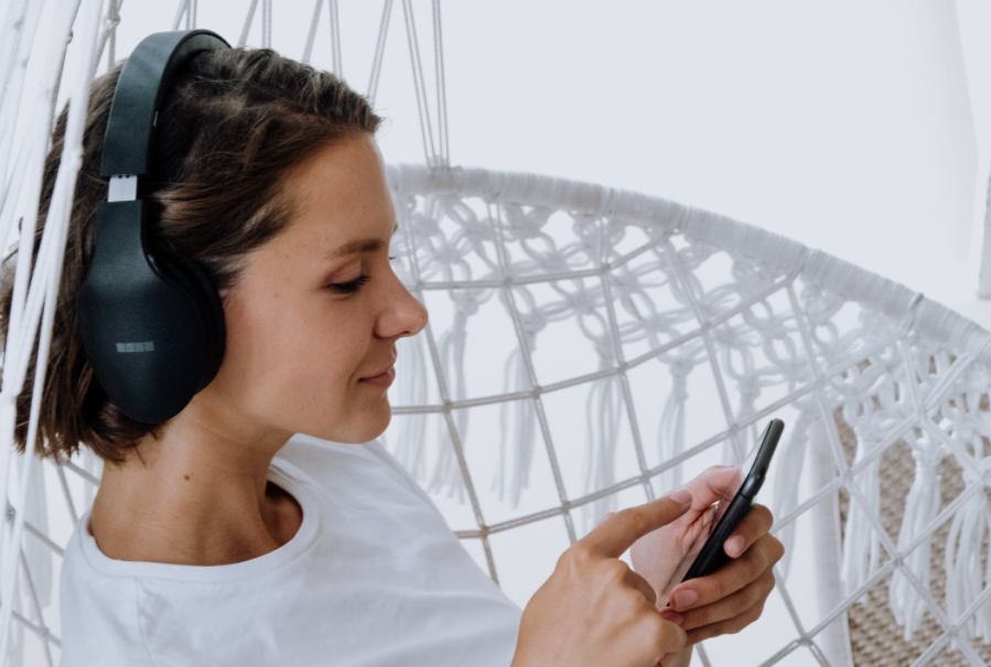 Mulher está sentada em uma poltrona branca de tiras. Ela está com fones nos ouvidos e segurando o celular enquanto relaxa.