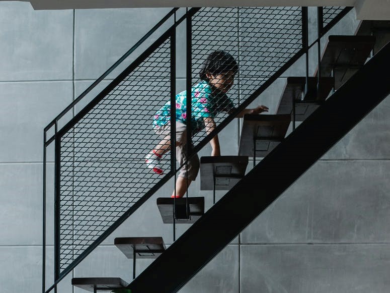Criança subindo as escadas. Para evitar acidente doméstico é preciso ter a vigilância de um adulto na hora que a criança for subir as escadas. A escadas são da cor preta e a parede é acinzentada.