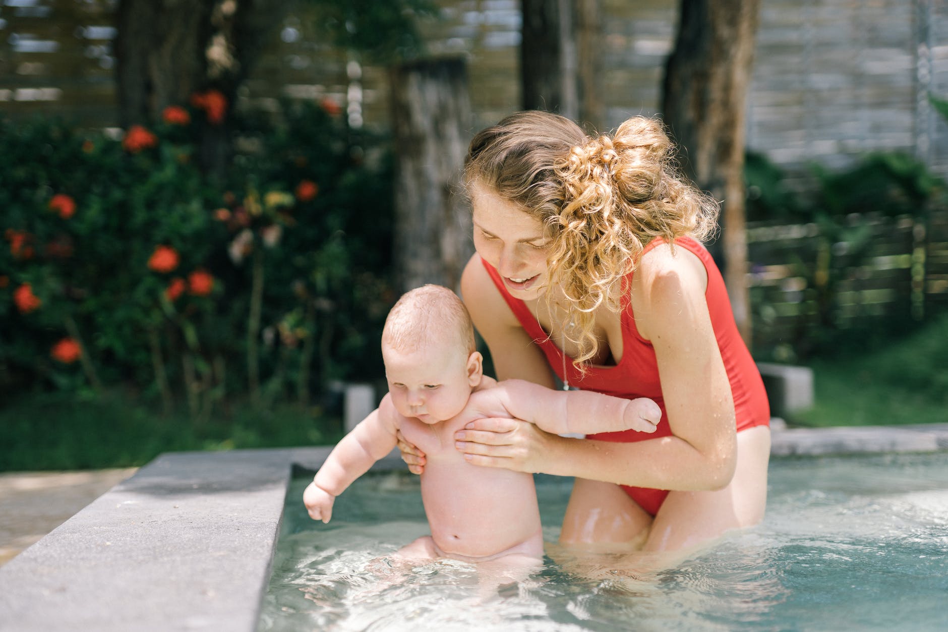 Mulher e bebê brancos brincam na piscina. A mulher tem o cabelo loiro e veste um maiô de cor vermelha, ela segura o bebê que aparenta ter meses de idade para prevenir acidente doméstico com a criança na água. Eles estão dentro de uma piscina rasa. Ao fundo há plantas e flores.