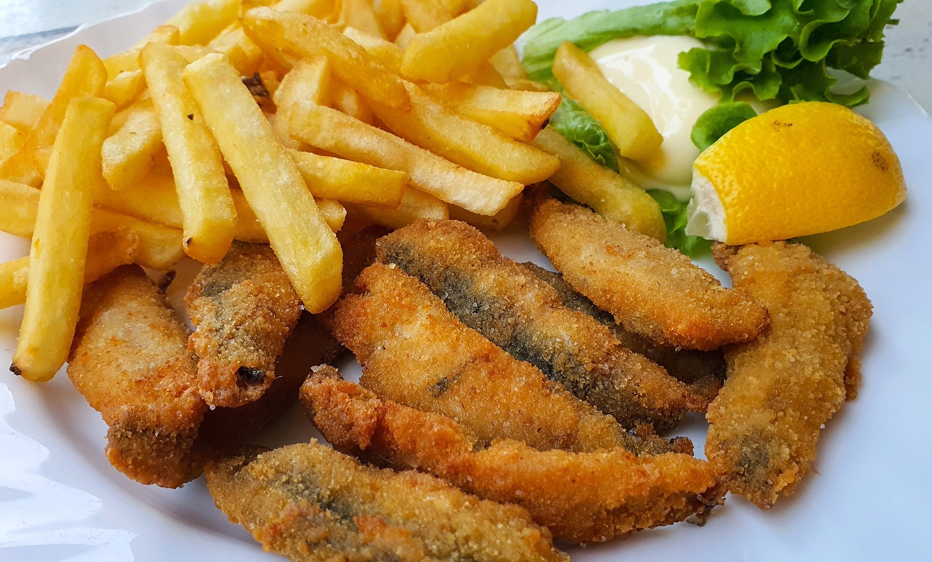 Sardinha frita com farinha é uma boa opção de receita saudável e com muito sabor. Várias sardinha empanadas com fubá estão no prato, há também batatinhas fritas, pedaço de limão e salada.