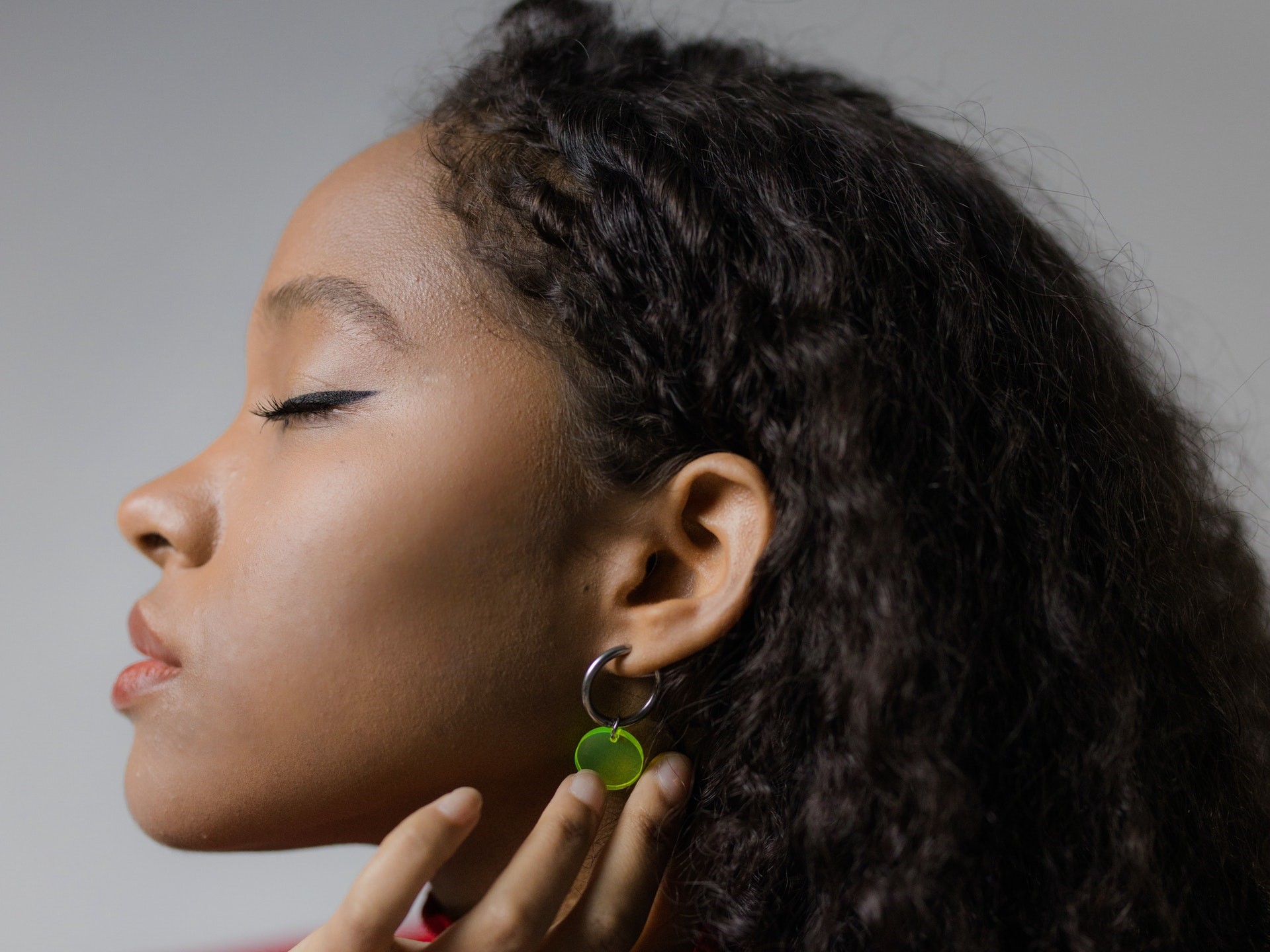 Mulher negra está de perfil. Ela usa um brinco de cor verde e está com os olhos fechados. O cabelo da mulher é longo e está solto, ingerir alimentos saudáveis ajudam a melhorar a saúde dos cabelos e auxiliam no crescimento capilar.