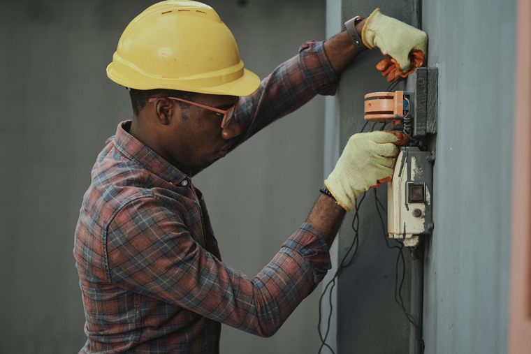 Homem negro usa capacete de proteção de cor amarela e veste camisa xadrez e luvas. Ele está mexendo no quadro de energia. A manutenção regular das instalações elétricas é essencial para evitar choques elétricos.