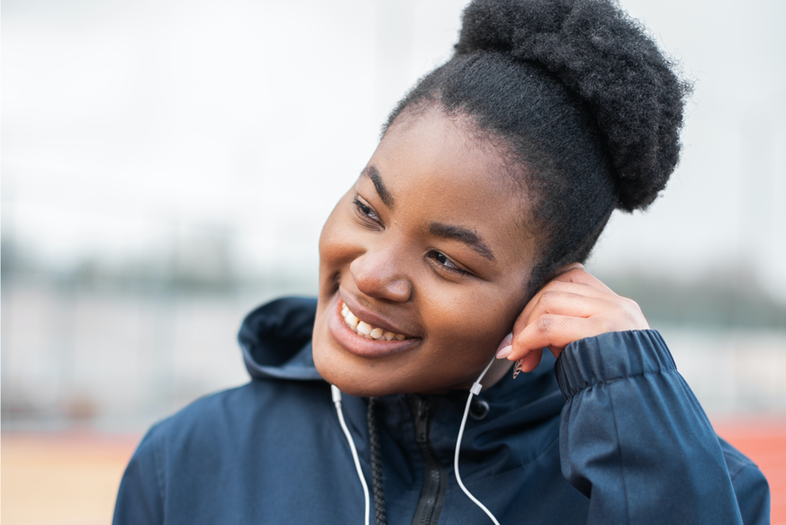 Mulher negra está sorrindo. Ela usa fones de ouvido e veste um moletom de cor azul. Para cuidar da saúde auditiva o indicado é não passar longos períodos com fones no ouvido.